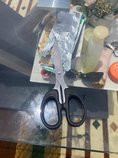The image shows a pair of black-handled scissors resting on a glass table. The scissors are positioned diagonally across the frame, with the blades pointing towards the left and the handles towards the right. The blades are made of metal and appear to be slightly worn.

On the table, to the left of the scissors, are several items. These include a clear bottle with a yellow cap, a pink tube of lotion, a small orange container, and some packets of medication. One of the packets has the text "3x1" visible. There are also some decorative flowers with pink and green hues, and a keychain with a black fob. The table surface is reflective, showing the items and the surrounding environment.

The setting appears to be indoors, likely a home or office, given the presence of personal items. The lighting suggests it could be daytime, but there are no strong indicators of the time of day or weather conditions. The surface of the table is somewhat dusty and scratched, indicating it's a well-used surface. The background shows a striped rug with brown and beige colors, and the edge of a dark surface, possibly another piece of furniture. No people are visible in the image. The overall impression is a candid shot of everyday objects on a table.