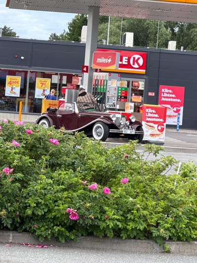 This media file captures a street scene at a gas station in Laagri alevik, Estonia. The main subjects are a vintage burgundy car and a woman standing near the entrance of the gas station. The car, a classic model, is parked partially obscured by a large, dense bush with pink flowers. A person, possibly the driver, is visible in the car wearing a plaid shirt.
The setting is a "K" branded gas station, indicated by the prominent "LEK" logo and "miles+" branding on the pillars. Several signs are visible, including red advertisements with the text "Kiire. Lihtne. Juustune." (Fast. Easy. Delicious.) and an offer for "Einedill!" (Meals!) at 7€ and 4.50€. The pumps are clearly visible, with numbers "3" and "4" indicated. A woman with blonde hair, wearing a blue top, is standing in front of the store entrance.
The weather appears to be overcast or at least not intensely sunny, with soft lighting. The presence of lush green foliage and blooming pink flowers suggests it is springtime or summer. The overall atmosphere is casual and everyday, with no indication of a special event. The visible license plate on the car reads "002 API". The scene is captured from a slightly elevated, tilted perspective.