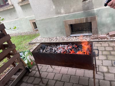 A close-up, eye-level outdoor shot captures a barbecue grill filled with glowing red coals and burning embers. To the left, a wooden stand holds a bag of charcoal and a grill brush. In the background, a building with light green and beige textured walls rises, featuring a metal grate resting against it and a recessed window. The ground is paved with gray tiles and scattered with small, light-colored pebbles and grass. Sparks are visible flying from the grill, indicating active burning. The scene appears to be in Freital, Germany, suggested by the architectural style.