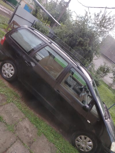 The image, taken in Rudzensk, Belarus, shows a black station wagon parked on a gravel and grass area next to a chain-link fence. The car is wet, indicating recent rain. The fence separates the parking area from a yard with trees and a building with a tiled roof. Part of a brick building and a wooden structure are visible in the background. The car's windows are reflective and show the surrounding environment. The overall scene suggests a residential or rural setting in the city.