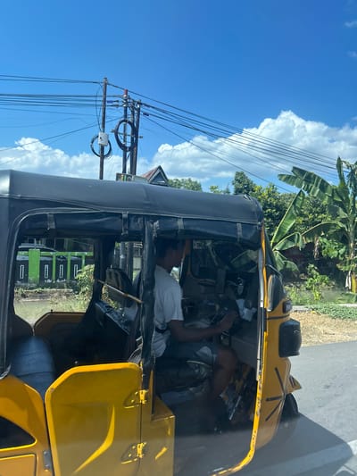 In Kalabbirang, Indonesia, a yellow and black auto-rickshaw, likely a Bajaj, is traveling on a paved road under a bright blue sky with scattered white clouds. The driver, an adult male wearing a white t-shirt and dark shorts, is seated behind the steering wheel. The auto-rickshaw has an open passenger compartment with a black fabric roof. In the background, there are utility poles with numerous wires, lush green vegetation including banana trees, and a building with a green fence. The scene appears to be in a semi-urban or rural setting, given the presence of both infrastructure and natural elements.
