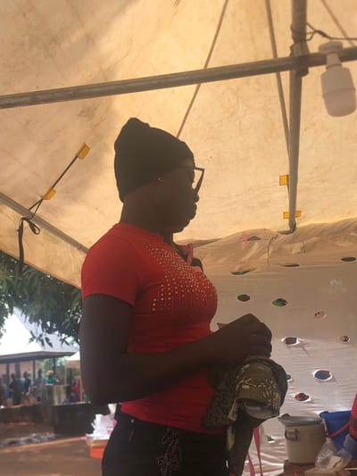 A woman is inside a tent in Nteje, Nigeria. She is wearing glasses, a black head covering, a red bedazzled shirt, and a black skirt. She holds a camouflage scarf in her hands. The tent has a light bulb fixture and support beams. Other people are visible outside of the tent.