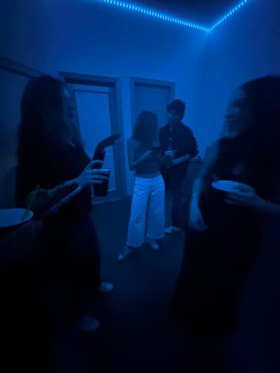 A room with blue lighting contains a group of people. One person in white pants is lying down, and another person in black clothing is sitting and playing the guitar. Two other figures appear, one above them holding a cup and another below holding a phone. A string of lights is visible along the top of the wall, and the room has dark shadows and low lighting.