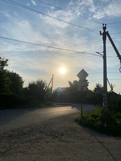 This image depicts a rural road intersection under a bright, hazy sky. The sun is high and slightly obscured by clouds, casting a warm, diffuse light. Overhead, power lines crisscross the frame, supported by utility poles. On the right side of the road, a prominent concrete utility pole stands, leaning slightly and reinforced by a diagonal support. Adjacent to it are two road signs: a large, empty diamond-shaped sign and a smaller square sign with a horizontal line. Further down the road, a blue square sign with a white pedestrian symbol is visible. The road itself is paved with asphalt and shows signs of wear, with gravel and patches of dirt on the shoulder. Lush green vegetation lines the sides of the road, with trees and bushes forming a dense backdrop. In the distance, rooftops of houses are visible, hinting at a small settlement. The overall impression is one of a quiet, semi-rural setting. The image was taken in Alamüdün, Kyrgyzstan.