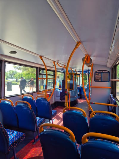 The image shows the interior of a bus, likely a Stagecoach bus, moving through South Cambridgeshire, United Kingdom. Rows of blue seats with patterned headrests line the aisle, with yellow handrails above. Through the bus window, two people are visible outside, one male in a dark jacket and cap walking past a green area and the second person is in a checkered skirt. An information panel is fixed to the wall at the front of the bus. The floor has a patterned red and blue carpet.