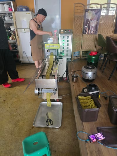 This media file captures an indoor scene, likely a food preparation area or a small workshop in Gumi-si, South Korea. The primary subject is a middle-aged man wearing a black cap, a black tank top, and a brown apron. He appears to be operating a noodle-making machine.

The machine itself is a prominent object, made of stainless steel with various controls and a conveyor belt system. Yellow noodles are visibly being extruded and processed by the machine. A spray bottle and a tray with scissors are also part of the equipment.

In the background, various objects are visible, including a large green water bottle with a red cap, what appears to be a portable cooler or storage container filled with more yellow noodles, a digital display panel with illuminated green lights, and what looks like a smartphone charging. The setting suggests a functional, possibly commercial, environment with a wooden floor and wall paneling. The overall impression is one of a busy workspace where food production is in progress. The lighting and angle of the shot indicate it was taken during daylight hours, possibly indoors with artificial lighting. No specific emotions are clearly discernible, but the man's posture suggests focused activity. There is some visible text on the machine and a label on the spray bottle, but it is not legible in detail.