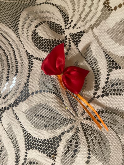 A red satin bow with two thin orange ribbon streamers is centrally placed on a white lace tablecloth. The lace has an intricate geometric pattern with open, diamond-shaped cutouts. The lighting creates shadows and highlights on the bow and the fabric, indicating an indoor setting. No people are visible in the image. The location in Konstantynów Łódzki, Poland is not discernible from the image itself.