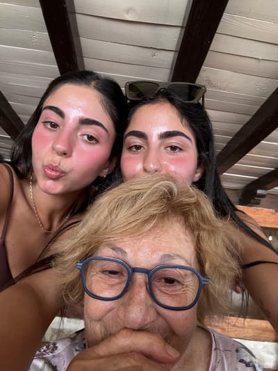 A close-up, selfie-style photograph shows three females positioned in tight proximity, purportedly taken in Cozumel, Mexico. The specific location within the city is not visually discernible. In the foreground, an older woman with short, wavy blonde hair is captured from the upper chest level. She wears blue-framed spectacles and a light-colored top with a floral pattern. Her right hand is raised, covering her mouth, as her eyes crinkle, indicating a smile or laughter. Behind her, two younger females with dark hair are visible. The female on the left, wearing a brown top and a delicate gold chain necklace, pouts her lips in a kissing gesture, and her cheeks show a distinct reddish flush. The female on the right has dark sunglasses perched on her head and her face is partially obscured by the older woman; her visible cheek also displays a reddish flush. The background consists of a light-colored, horizontally slatted wooden ceiling or roof structure, intersected by darker wooden support beams, consistent with an outdoor covered area receiving bright daylight.