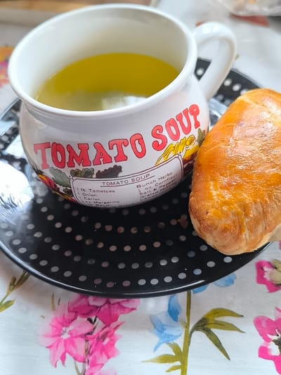 A close-up, high-angle shot shows a white ceramic mug with red lettering that reads "TOMATO SOUP" on a patterned black plate. The mug contains a yellow liquid, likely soup. Next to the mug, on the right side of the frame, is a golden-brown baked good, possibly a pastry or bread roll. The plate rests on a white tablecloth with a floral pattern of pink, blue, and green flowers. The scene appears to be indoors, possibly in a kitchen or dining area. No specific location in Žiaukos, Lithuania, can be determined from the image.