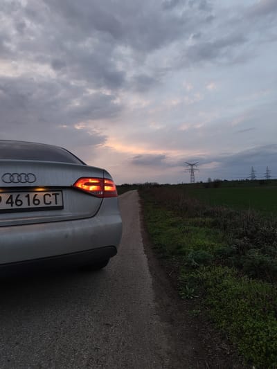 The image captures the rear quarter view of a silver Audi sedan positioned on the left side of a narrow, asphalt-paved road in Gorni Tsibar, Bulgaria, during twilight conditions. The vehicle's Bulgarian license plate, displaying "P 4616 CT", is clearly visible, with its frame partially obscuring the top of the 'P'. The Audi's red taillights are illuminated. To the immediate right of the road, a dirt shoulder transitions into dense, low-lying green vegetation and uncultivated scrubland. Beyond this vegetation, a large green field stretches into the distance. In the far background, multiple large metal electricity transmission towers (pylons) stand against the horizon, supporting power lines that traverse the landscape. The sky is predominantly covered with heavy, textured grey clouds, with a segment near the horizon exhibiting lighter hues and faint orange-pink tints, suggesting the setting sun or twilight glow. No individuals or specific actions are discernible within the frame.