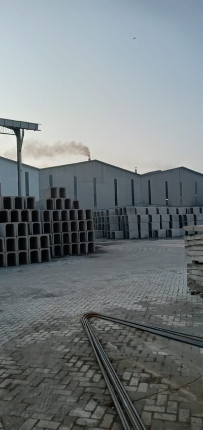 The image shows a concrete manufacturing facility in Sumput, Indonesia. Stacks of concrete culverts and flat slabs are visible in the foreground and midground, along with a coil of rebar resting on a brick-paved yard. In the background, multiple prefabricated metal buildings with smoke emanating from one chimney are present. An overhead crane structure is visible on the left, and a single bird is in the sky.