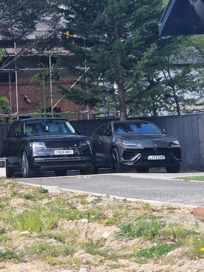The image captures two luxury black SUVs parked outdoors in Witton Gilbert, United Kingdom, under bright daylight, suggesting a clear and sunny day. The scene shows the vehicles positioned on what appears to be a paved area or driveway, adjacent to a sidewalk and a strip of unkempt ground.
The vehicle on the right is a black Lamborghini Urus, clearly displaying the UK registration plate "LJ72 CCO". Its aggressive front grille and styling are prominent. The vehicle on the left is a black Range Rover, identifiable by the "RANGE ROVER" text across its bonnet, and bearing the UK registration plate "DN24 MXT".
In the background, dense green foliage from trees dominates, partially obscuring a brick building. Scaffolding is visible against the brickwork of the building, suggesting ongoing construction or renovation. A dark wooden fence or building facade is situated directly behind the Lamborghini.
The foreground and right side of the image show a paved sidewalk or path bordering a strip of dry earth mixed with green grass and small weeds, some bearing tiny yellow flowers. No people are visible, and the primary activity captured is the stationary presence of the two vehicles.
Visible text includes the two distinct UK license plates: "LJ72 CCO" on the Lamborghini Urus and "DN24 MXT" on the Range Rover. The "RANGE ROVER" branding is also clearly seen on the second vehicle. A partial, blurry number "2" can be faintly seen in the bottom right corner of the original image. The overall setting appears to be a residential or semi-urban area.