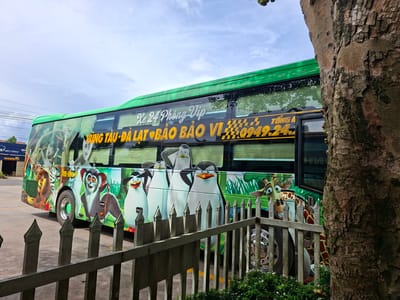 A brightly painted green bus, adorned with cartoon characters from the movie Madagascar, is parked in a lot. The bus features images of penguins, a lemur, a lion, a zebra, and a giraffe. Text on the side of the bus reads "Xe 24 Phong Vip Vung Tau - Da Lat - Bao Bao Vi" and includes a phone number. The bus is partially obscured by a large tree trunk on the right side of the frame and a metal fence in the foreground. The ground is paved, and a blue sky with scattered clouds is visible above. In the background, a building with a "MAXILITE" sign is present. The location is identified as Tân Phú 2, Vietnam.
