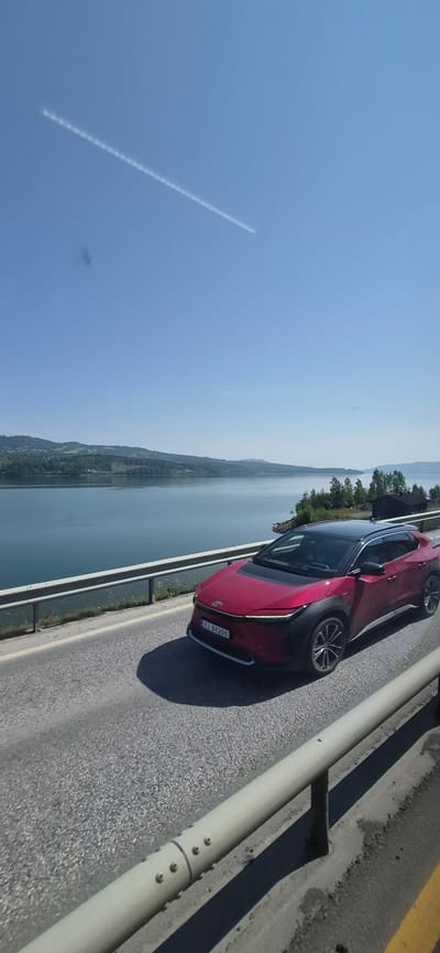 An eye-level, vertical, medium shot captures a crimson Toyota bZ4X electric SUV driving on a paved road alongside a large body of water, with a forested, hilly landscape in the background. The car, facing right, has a black roof and black trim around the wheel wells and lower body. Its license plate, "EJ 84200", is visible on the front. The road is gray asphalt with a white stripe on the right edge. A gray metal guardrail runs along the left side of the road, separating it from a drop-off toward the water. The water is a calm, deep blue. The background features green hills covered in trees, with some houses and structures visible. The sky is a clear, bright blue with a faint white contrail from an airplane stretching across it from the upper left to the middle right. The overall lighting suggests a sunny day. The location is Vingrom, Norway.