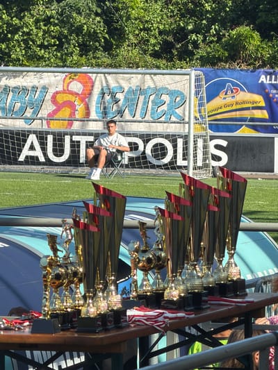 Here is a forensic description of the image:
The image depicts an outdoor scene, with a soccer field, trees, banners, and trophies. In the background, there's a green field and trees on the left side. A banner is visible, with text "ALL" and "Groupe Guy Rollinger" and an "A" logo. Adjacent is a net with the words "AUT POLIS" vertically printed. A man sits on a chair behind the net. In the foreground, there are multiple trophies arranged on a table. Red and white striped ribbons decorate some of the trophies. The location appears to be in Fentange, Luxembourg, based on the banner information and OCR text.
