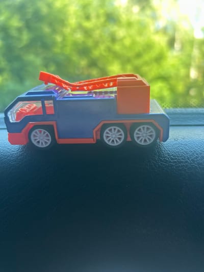 A toy truck is photographed close-up, with its wheels facing the viewer. The truck is primarily blue and orange, with white wheels. The background is blurred and green, suggesting foliage outdoors. The truck appears to be resting on a dark, textured surface, possibly the dashboard of a vehicle. The lighting suggests it is daytime, with the sun possibly shining through a window, casting bright highlights on the background. The image is oriented vertically. There is no visible text.