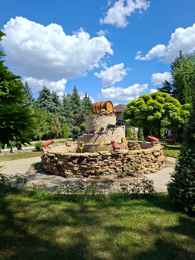 A tiered fountain constructed from rough-hewn stones is the central focus of this outdoor scene. The fountain is adorned with decorative elements including clay amphorae, wine bottles, and a wooden barrel at its apex. Two glasses of what appears to be white wine are placed on the stone structure, suggesting a wine-themed setting. The fountain is surrounded by a paved walkway and a manicured lawn, with various trees and shrubs providing a lush green backdrop. In the background, a building with a gabled roof is visible, hinting at a larger estate or establishment. The sky above is a vibrant blue, dotted with fluffy white clouds, indicating a pleasant, sunny day. The overall impression is that of a picturesque outdoor space, likely designed for leisure or to highlight a connection to viticulture. This scene is located in Milestii Mici, Moldova, a region renowned for its extensive wine cellars and winemaking traditions.