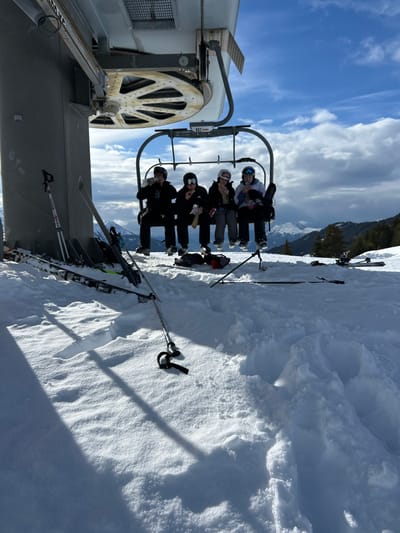This image depicts a snowy mountain scene in Villard-sur-Doron, France, under a partly cloudy blue sky. Four individuals are seated on a suspended grey ski lift chair, positioned directly below the large metallic wheel mechanism of a ski lift pylon visible on the left. All four subjects are wearing winter ski or snowboard attire, including helmets or beanies and goggles.
From left to right, the first subject, in a black jacket and dark helmet, holds a container to their mouth, appearing to drink. The second subject, also in a black jacket and dark helmet with goggles pushed up, holds a light-colored wrapped item, possibly a snack or sandwich. The third subject, wearing a dark jacket and a light-colored beanie with goggles pushed up, similarly holds a light-colored wrapped item. The fourth subject, in a white jacket and white helmet with goggles covering their eyes, holds a container to their mouth, appearing to drink. Two dark bags rest in the snow directly below the chair.
The ground is covered in deep, white snow, exhibiting varied textures and casting long shadows from the bright sunlight. Several pairs of skis and ski poles are visible on the snow, some propped against the pylon base on the left, and others lying flat in the foreground and to the right. A single ski pole lies horizontally in the central foreground snow. Distant snow-covered mountains and evergreen trees are visible under the horizon line, complementing the high-altitude ski resort environment. The ski lift chair bears the number "802 DURON" on a small label.