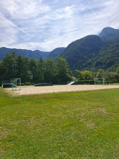 The image depicts a sunny outdoor recreational area located in Obertraun, Austria. In the foreground, a lush green lawn occupies the lower portion of the frame. The midground features a sand court with a small soccer goal on the left and a ramp or skate feature on the left side of the sand. Beyond the sand court, a line of mature trees forms a dense green backdrop. In the distance, imposing mountains rise, their slopes covered in dark green forests, under a bright blue sky with wispy white clouds. The scene conveys a sense of a peaceful, natural environment suitable for outdoor activities.