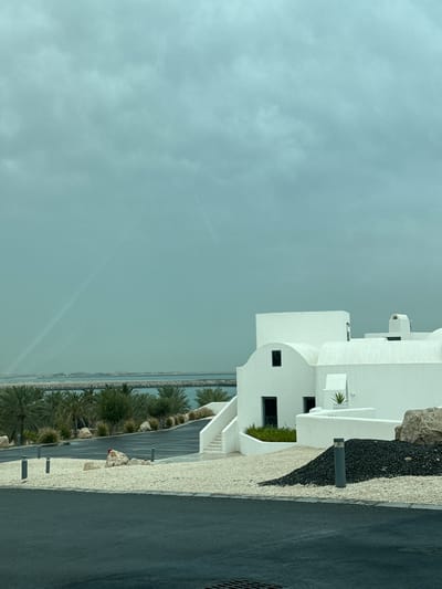 The image shows a modern, white building adjacent to a paved area in Ghadeer Al Tayr, United Arab Emirates. The building features rounded architectural elements, multiple levels, and small windows. Along the pavement, there's a landscaping design incorporating black and white gravel, interspersed with rocks and evenly spaced light posts. Beyond the pavement and landscaping, a row of palm trees separates the built environment from a body of water, possibly the Persian Gulf, under a cloudy sky. The water and sky blend into a homogenous grayish hue. No people are visible in the frame.
