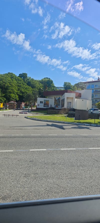 The image captures a street scene in Vostok, Russia, under a blue sky with scattered clouds. The foreground is dominated by a grey asphalt road with a white dashed center line.

On the far side of the road, there is a low building with white walls and a red roof. The building features an outdoor mural or artwork on one of its walls. In front of the building, there is a small patch of green grass.

Behind the building, trees are seen. A light-colored vehicle is parked near the building. Further to the left, there is a gatehouse with a barrier and a "STOP" sign. The scene appears to be taken from inside a car.