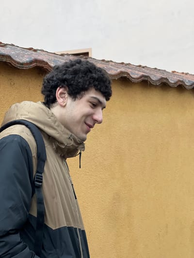 A young adult male, with dark, curly hair, is positioned in the foreground, facing right with his head slightly tilted downwards. He exhibits a faint smile, directing his gaze to his lower right. He wears a two-tone jacket, beige on the upper section and dark (black or navy) on the lower half, with what appears to be a dark backpack strap visible over his right shoulder. Small water droplets or stains are visible on the beige portion of his jacket. The background features a textured wall painted a golden ochre color, above which a terracotta-tiled roofline is visible. Water droplets are present along the edges of the roof tiles. Above the roofline, an overcast, light grey sky is visible, with a small, indistinct light-colored structure appearing in the upper left background. The scene is located in Meistratzheim, France.