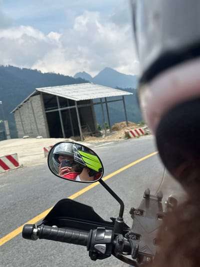 An image captured from the first-person perspective of a motorcycle rider depicts a scenic mountainous road. The photo is oriented ninety degrees counter-clockwise, showing the sky and mountains on the left and the road and motorcycle parts on the right.
When viewed upright, the bottom right of the frame features the motorcycle's black handlebar with textured grips and control switches, and a phone mount attached near the windshield. A circular rearview mirror in the center-right prominently reflects the rider. The rider is wearing a light-colored helmet, a bright yellow-green high-visibility vest over a red shirt, and is holding a smartphone to take the picture, visible in the mirror's reflection.
The paved road occupies the central and left portions of the frame, marked by a solid yellow line running along its edge. Red and white striped barriers or hazard markers are positioned along the roadside, indicating a potentially steep drop-off or a construction area. Beyond the road and barriers, a rocky, dirt embankment with sparse vegetation leads to a more developed background.
In the mid-ground, an open-sided building with a corrugated metal roof and exposed metal beams stands, possibly a shelter or an unfinished structure. The far background reveals majestic mountains under a partly cloudy sky, characteristic of a high-altitude or rugged terrain. The scene suggests a journey through a remote, possibly rural, mountainous region during daylight hours with fair weather.