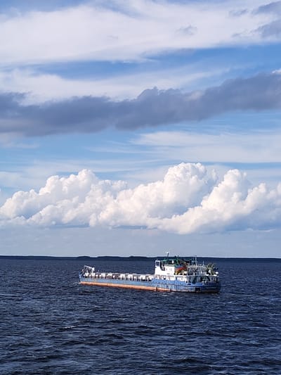A blue and orange barge travels on a dark, choppy body of water. The barge has a white superstructure with various equipment visible. The water extends to a distant, dark shoreline under a sky filled with a mix of white and grey clouds. The scene appears to be the city of Myaksa, Russia.