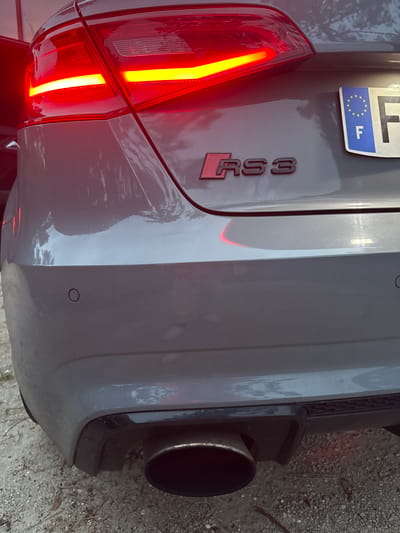 A detailed rear view of a grey Audi RS3, parked on a light gravel surface in Lège-Cap-Ferret, France, is captured under ambient daylight. The vehicle's red LED taillights are illuminated, featuring distinct horizontal yellow indicator strips. Centered on the trunk lid is a prominent red and black "RS3" emblem. To the right, a partially visible French license plate shows the blue European band with the letter "F" and a circle of stars, with the main plate beginning with "F". The lower bumper includes a black textured diffuser housing a single, large, dark metallic exhaust pipe on the left side, with another exhaust likely present on the right (not fully visible). A circular parking sensor is clearly visible on the lower left portion of the bumper, with another partially visible on the right. Reflections on the car's paintwork suggest an outdoor environment. No people or active interactions are visible in the scene.