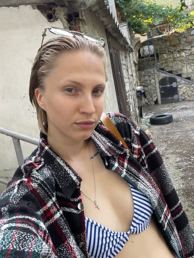 This is a slightly rotated, close-up selfie of a woman with fair skin and blonde hair, wearing a blue and white striped bikini top and an open plaid jacket. She is wearing glasses perched on her head. A thin silver necklace with a small cross pendant is visible. The background appears to be an outdoor, possibly slightly dilapidated, area with stone walls, a metal railing, and some greenery. The lighting suggests it is daytime, with natural light illuminating the scene. The woman's expression is neutral, looking directly at the camera. The background details include a black tire mounted on a wall and some rubble. The overall impression is a casual, personal photograph taken in an outdoor setting. The provided location context is Гаспринский поселковый совет, Ukraine.