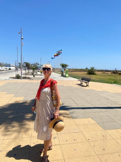 A woman stands on a paved walkway under a clear blue sky. She is wearing a light-colored, tiered dress, a red scarf around her neck, sunglasses, and sandals. She carries a straw bag over her right shoulder, a bottle in her left hand, and a straw hat with a black ribbon in her right hand. The walkway is made of light brown and grey square tiles, with shadows cast by lampposts and palm trees. In the background, several flags are visible on poles, and a bench sits on the walkway. Beyond the walkway, there is a grassy area with a wooden fence, palm trees, and a beach in the distance. The scene appears to be in Conil de la Frontera, Spain, likely near the coast, given the flags and beach visible.