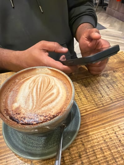 The image shows a person holding a smartphone taking a picture of a cappuccino, likely sitting at a table in a cafe or similar establishment. The cappuccino is in a grey ceramic mug with latte art, resting on a matching saucer with a spoon. The table appears to be made of textured wood.
Location: This detail is not discernible from the provided image and would be impossible to ascertain without additional information.