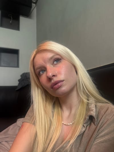 A blonde female adult is depicted in an indoor setting, with her head slightly tilted upwards and to the right, and her blue eyes directed similarly. Her facial skin exhibits localized redness on the cheeks and nose. She wears a light-colored, possibly beige or light brown, collared shirt with a subtle sheen or texture, and a white pearl necklace. A small reddish mark is visible on her left upper arm. The background consists of a light grey wall featuring two dark, empty square recesses and a dark object, likely a speaker or light fixture, mounted above them. A dark sofa or upholstered seating is partially visible to the left. The specific location within Namyangju-si, South Korea, cannot be determined from the visual details present in the image.