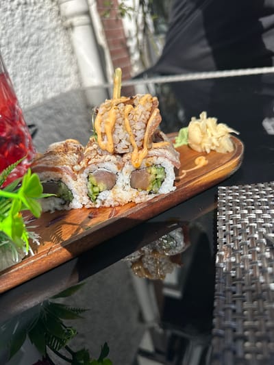 The image shows a wooden serving board with sushi rolls, garnished with pickled ginger, wasabi, and sauce. The sushi appears to be on a reflective surface, likely a table. The backdrop includes elements of an outdoor setting with white concrete and black decorative metal work which is common to many establishments in Rotenburg an der Fulda, Germany. No people are visible.
