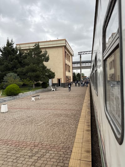 The image depicts a train station platform in Jizzakh, Uzbekistan, during overcast weather. A train is parallel to the platform on the right. A group of people is congregated further down the platform, possibly waiting for or disembarking from the train. The platform is constructed of red and gray pavers with a yellow safety strip near the train tracks. Adjacent to the platform is a landscaped area with trees and benches. A multi-story building is present, partially visible behind the trees.