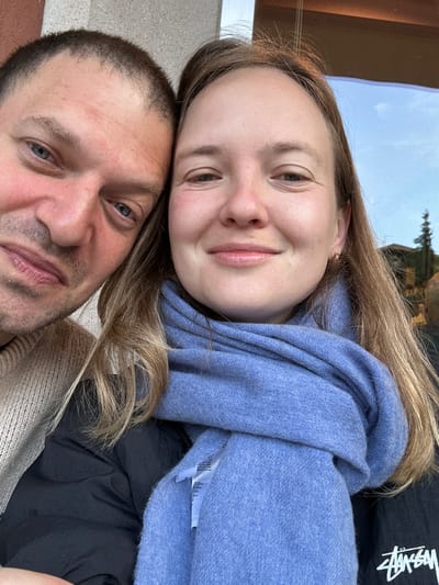 A close-up portrait depicts a male and female adult positioned closely together, looking at the camera and smiling. The male, on the left, has short dark hair, blue eyes, and light facial hair, wearing a light beige knit sweater. The female, on the right, has long light brown hair, a gold hoop earring in her right ear, and is wearing a black puffer jacket with a light blue knitted scarf wrapped around her neck. The background behind the male is a light-colored, textured wall. Behind the female, a window reflects a blue sky with some clouds, the tops of evergreen trees, and a distant building with a reddish roof, indicative of an outdoor or indoor setting with an external view in Longone al Segrino, Italy.