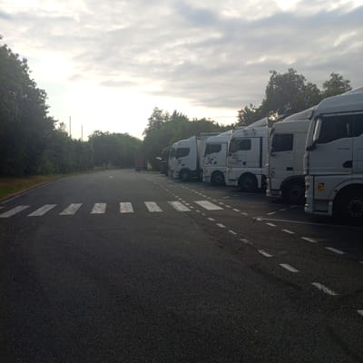 The image shows a row of parked semi-trucks on the side of a road. The trucks are white and appear to be part of a logistics company, as indicated by logos on their sides. The road has white markings, including a pedestrian crossing in the foreground. Trees line the left side of the road, and the sky is cloudy. There are no people visible in the image. The scene appears to be a rest stop or parking area for commercial vehicles. The location is identified as being in Saint-Augustin-des-Bois, France, though this cannot be visually confirmed from the image alone.