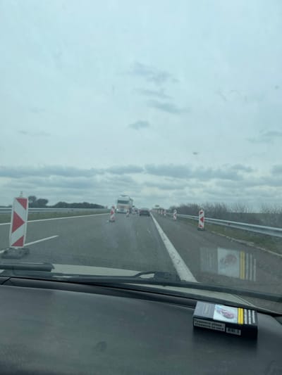 The image depicts a forensic view from inside a vehicle traveling on a highway in Vinarsko, Bulgaria, under an overcast sky. In the immediate foreground, a black dashboard is visible, with a pack of "TENOR" cigarettes resting on it. The cigarette pack displays Bulgarian text indicating a health warning and features a graphic image. Through the windshield, the scene ahead shows a multi-lane asphalt road with visible white lane markings. Road maintenance or construction is indicated by numerous red and white striped barriers placed along both sides of the lanes, directing traffic. In the midground, a white semi-truck is observed ahead, followed by several passenger vehicles. The distant background reveals sparse, likely deciduous vegetation along the roadside and a metal guardrail on the right. No individuals are explicitly visible. The overall environment suggests a cool, cloudy day with ongoing road work impacting traffic flow.