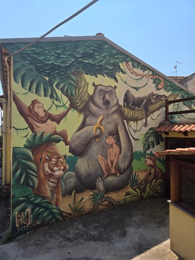 The image depicts a vibrant mural painted on the side of a building, likely in Viterbo, Italy, given the style of architecture and the presence of a street sign labeled "20". The mural features characters from Rudyard Kipling's "The Jungle Book," including Mowgli, Baloo the bear, Bagheera the panther, Kaa the snake, and Shere Khan the tiger, along with a monkey. Mowgli is shown sitting with Baloo, who is holding a banana. Bagheera is lounging on a branch above, and Shere Khan is depicted in the lower left corner. The mural is rendered in a colorful, somewhat whimsical style, with lush greenery and a clear blue sky as the backdrop. The building itself has a terracotta-tiled roof and a wooden structure attached to its side. The ground in front of the mural is paved with concrete, with some patches of shadow from the sunlight. The mural is signed with "RAJE MES" and dated "2019."