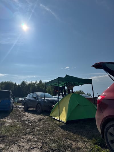 The image depicts an outdoor scene, likely a campsite, with several vehicles and camping equipment. The ground is muddy and appears to be dirt with some patches of grass. In the foreground, a green and yellow tent is pitched, and a portion of a red car with its trunk open is visible on the right. Behind the tent, a dark grey car is parked. A green canopy tent is set up over some items, and a person is partially visible standing under it. To the left, a blue van is parked. In the background, a dense line of evergreen trees forms a treeline against a bright blue sky with scattered clouds. The sun is shining brightly in the upper left corner of the image, creating lens flare. The scene suggests a temporary outdoor gathering or event. The presence of multiple vehicles and camping gear implies a recreational or temporary living situation. The environment is rural or semi-rural, given the treeline and the unpaved, muddy terrain.