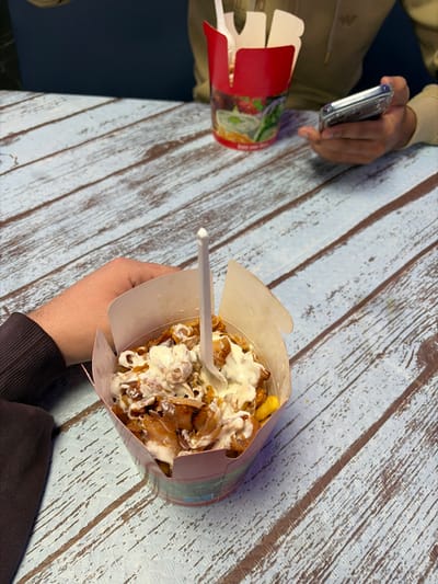 The scene is an indoor dining area in the city of Grau I Platja, Spain, featuring two individuals seated at a light blue, distressed wooden table. In the foreground, a person's left arm, clad in a dark brown long-sleeved garment, holds a light-colored paper container. This container is filled with a meal composed of french fries, pieces of cooked meat, and a generous topping of white sauce; a white plastic fork is embedded in the food. In the background, a second individual, wearing a light brown long-sleeved top, holds a silver smartphone enclosed in a clear protective case with both hands. A red paper take-out container, decorated with salad imagery and containing a white plastic fork and an indistinct meal, sits on the table directly in front of this second person. The background is an unadorned dark blue surface.