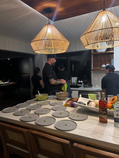 The image shows a kitchen island in a modern, upscale home, likely located in a residential area near the coast in Playa Herradura, Costa Rica. The scene depicts a group of people preparing food, likely for a party or gathering. Two men, wearing black shirts, stand near the island, while a woman in a black shirt stands to the left of them. There are multiple plates on the counter, many of them with small pieces of green food on them. There are also bowls with guacamole and various other ingredients, including fruits and a bottle of alcohol. Two wicker-style lights hang from the ceiling above the island. The room is illuminated by a combination of interior lighting and natural light coming in from outside the large windows. The windows offer a glimpse of the surrounding lush tropical environment. The atmosphere is one of casual conviviality and preparation.