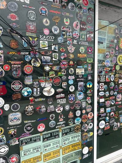 The image shows a close-up, eye-level view of a window covered in numerous stickers. The stickers are varied in size, shape, and design, with many featuring motorcycle-related imagery, logos, and text, some in Thai. Some stickers appear to be for specific clubs or events. Several black electrical wires are strung across the stickers, obscuring parts of the display. Behind the stickers, the glass reflects some of the surrounding environment, including what appears to be other windows and possibly buildings. Several license plates with Thai characters are also visible through the glass, suggesting the location is likely Thailand. The overall impression is of a heavily decorated window, possibly of a shop or a private residence. The presence of Thai license plates and the style of some of the stickers suggest it could be in a location like Ban Thai–Changlon, Thailand. No people are clearly visible.