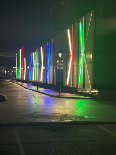 The image depicts a street scene at night in Ilidža, Bosnia and Herzegovina. A modern building facade is illuminated by a series of vertical LED lights in red, green, and blue. The lights reflect on the wet asphalt of the street and sidewalk, creating colorful patches of light. Lampposts are visible along the sidewalk. A "no entry" sign is mounted on a pole near the building. The overall atmosphere is urban and contemporary, with the vibrant lighting contrasting with the dark night sky.