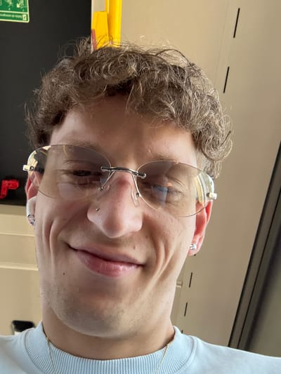 A close-up, selfie-style, waist-up shot shows a light-skinned male with curly brown hair and a nose piercing. He is wearing rimless sunglasses with a silver frame, a light blue crew neck sweatshirt, and a silver chain necklace. He has a small silver stud earring in his left ear and a larger, diamond-like stud earring in his right ear. He is smiling with his mouth closed. The background appears to be indoors with a yellow and black panel visible on the left, and a light beige wall with dark vertical lines on the right. There is a reflection of what appears to be a roadway with trees in his sunglasses. This image was taken in Móstoles, Spain.