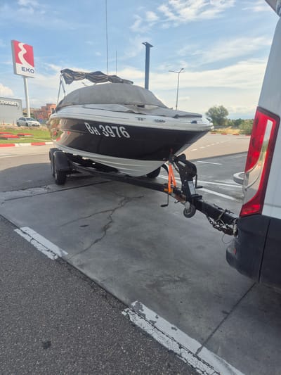 A white and black motorboat is parked on a trailer on a paved surface. The boat is covered with a grey tarp, and the registration number "BH 3976" is visible on its side. The trailer is attached to a white van, which is partially visible on the right side of the image. In the background, there is an EKO gas station sign and a McDonald's building. A white car is parked near the McDonald's. The sky is blue with some clouds. The scene is located in Chuchuligovo, Bulgaria.