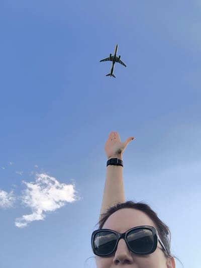 A person is photographed from below, looking up at a clear blue sky. An airplane is visible in the upper portion of the frame, flying against the blue sky. The person's arm is extended upwards, with their hand open as if reaching for the airplane. The person is wearing sunglasses and a dark watch on their wrist. A few wispy white clouds are present in the sky, towards the left side of the image. The scene is outdoors.