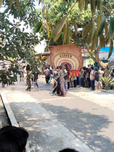The image depicts a scene in Botok, Indonesia, likely a street during a cultural event or parade. A large, ornate fan-like structure with the word "GATOEK" printed on it is being carried by a group of people, some of whom are dressed in traditional attire. Several other individuals, including men, women, and children, are gathered around, observing the procession or participating in the event. The environment appears to be outdoors, with trees, buildings, and a paved street visible. The lighting suggests it is daytime, with sunlight casting shadows on the ground. The overall atmosphere seems festive and communal.