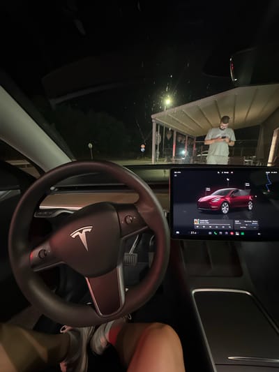 The image captures the interior of a Tesla vehicle at night, situated in Zagore, Bulgaria. The view is from the driver's seat, looking forward through the windshield. The dark-toned steering wheel, featuring the Tesla logo, dominates the lower left-center foreground. To the right, the illuminated central touchscreen displays a red Tesla vehicle graphic with an "Open Frunk" indicator. Below this, media controls are visible, showing "Family Affair" by Mary J Blige playing for 10 seconds. The screen also displays navigation details to "Killarney," indicating a remaining travel time of 1 hour and 44 minutes, a distance of 109 kilometers, and a battery charge of 53%. The time is 22:08, and the cabin temperature is set to 21.0°C, with an external temperature of 21°C.
In the bottom frame, the driver's lower legs are visible, clad in white sneakers with red detailing and white socks.
Through the windshield, an external night scene is illuminated by artificial light. A bright street light is visible to the left, casting light onto a distant background. A light-colored, open-sided shelter or building structure is prominent. Underneath this structure stands an adult male with a beard, dressed in a light-colored t-shirt and shorts, focused on a smartphone in his hands. The windshield exhibits faint reflections and what appear to be water droplets. In the far distance to the left, two circular road signs are faintly discernible, and dark foliage is present.