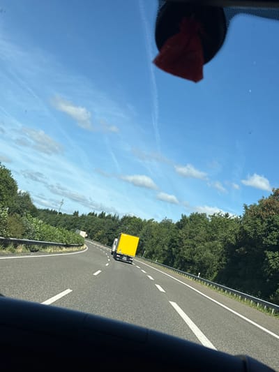 The image is taken from inside a vehicle, looking out through the windshield at a highway in Masburg, Germany. The sky is clear blue with scattered white clouds. A red air freshener hangs from the rearview mirror. The road ahead curves to the left, bordered by a metal guardrail and dense green trees. A yellow truck is visible in the distance, driving on the right side of the road. Another truck, mostly obscured by the yellow truck, is also present. The road surface is asphalt with white lane markings. The overall scene depicts a sunny day on a rural highway.